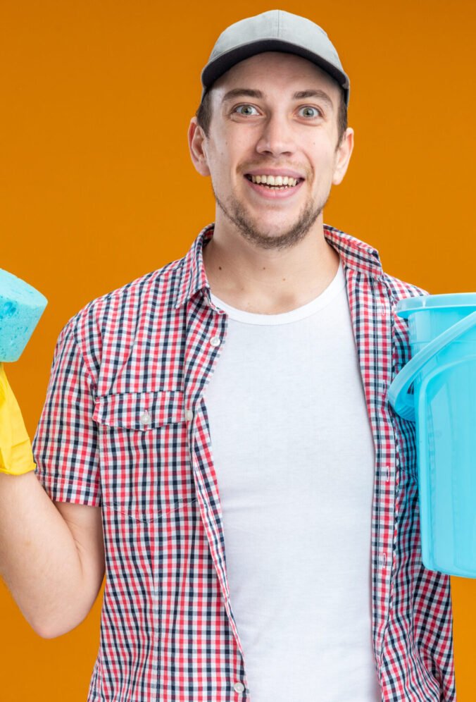 smiling young guy cleaner wearing cap with gloves holding sponge with bucket isolated on orange background